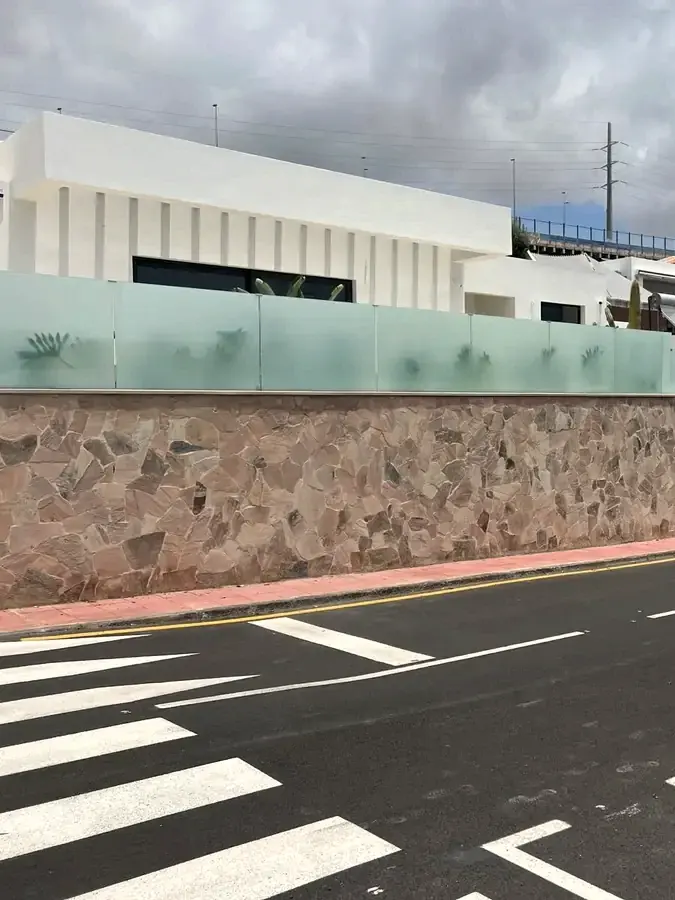 Muro de piedra con barandilla de vidrio y vial de acceso a una urbanización